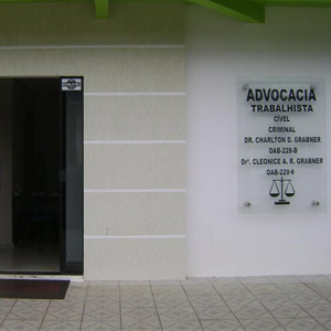 Na parede de cor neutra do escritório de Vilhena/RO, a placa de vidro evidencia a fachada da banca. 